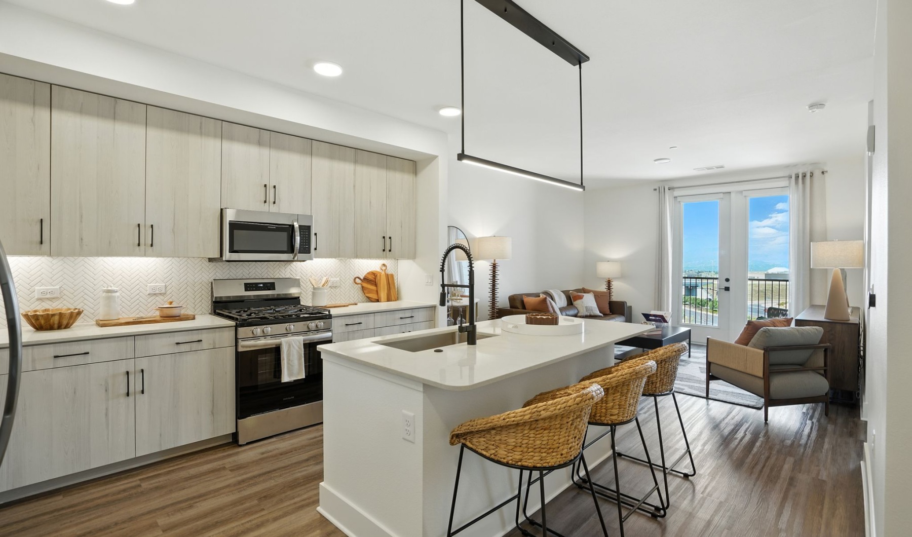 Modern open-concept kitchen and living area featuring sleek cabinetry, a spacious island with chic bar stools, and a cozy seating space with a beautiful outdoor view at The Aven in Colorado Springs